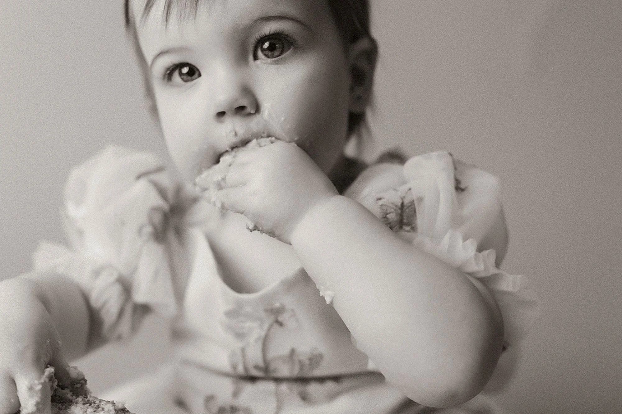 Black and white close-up of a baby eating cake with both hands, frosting on her face, candid moment from a simple cake smash photography session
