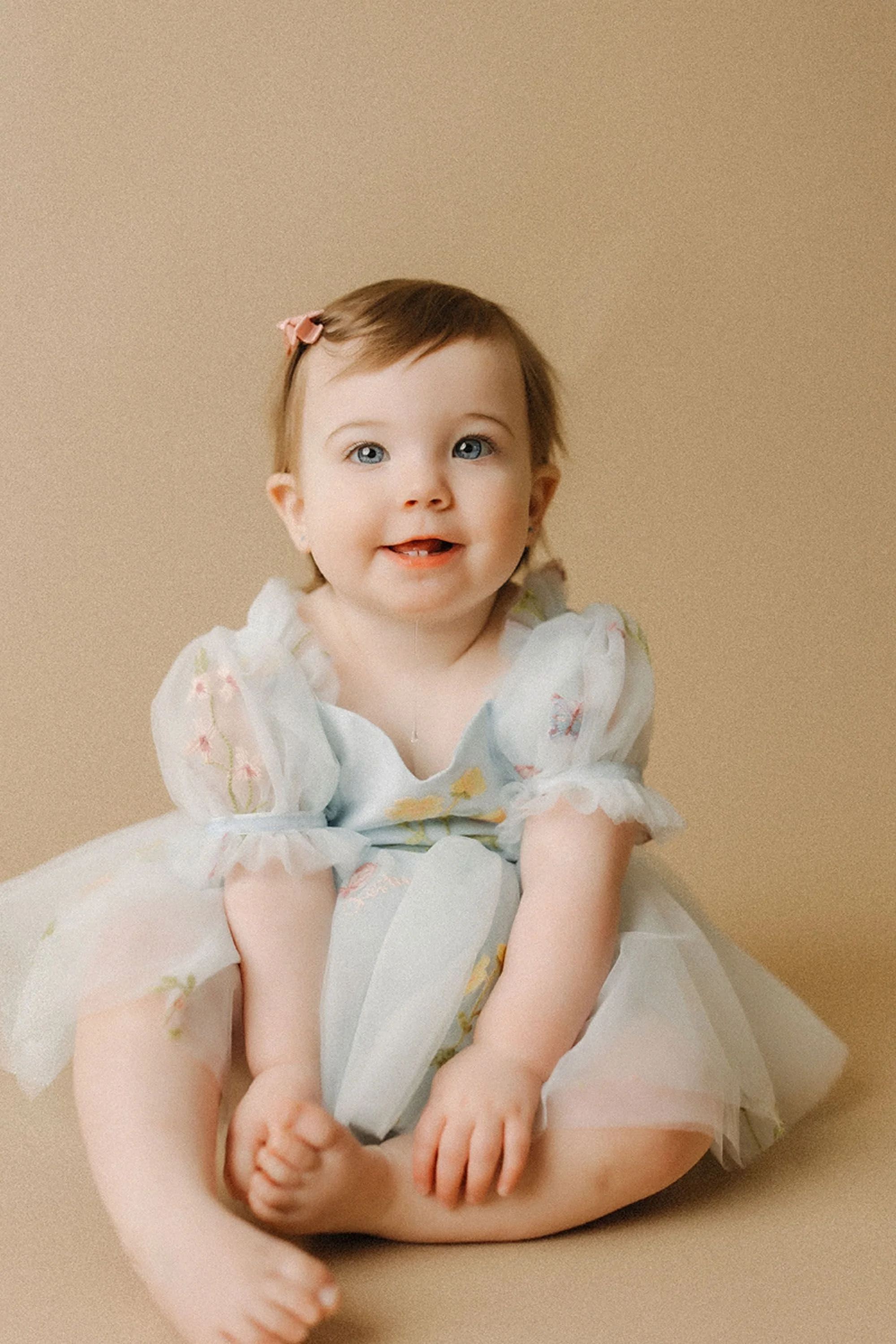 Simple Cake Smash Photos of a smiling baby girl in a delicate blue floral dress, sitting barefoot in a minimalist studio and looking happily toward the camera before her first birthday celebration.