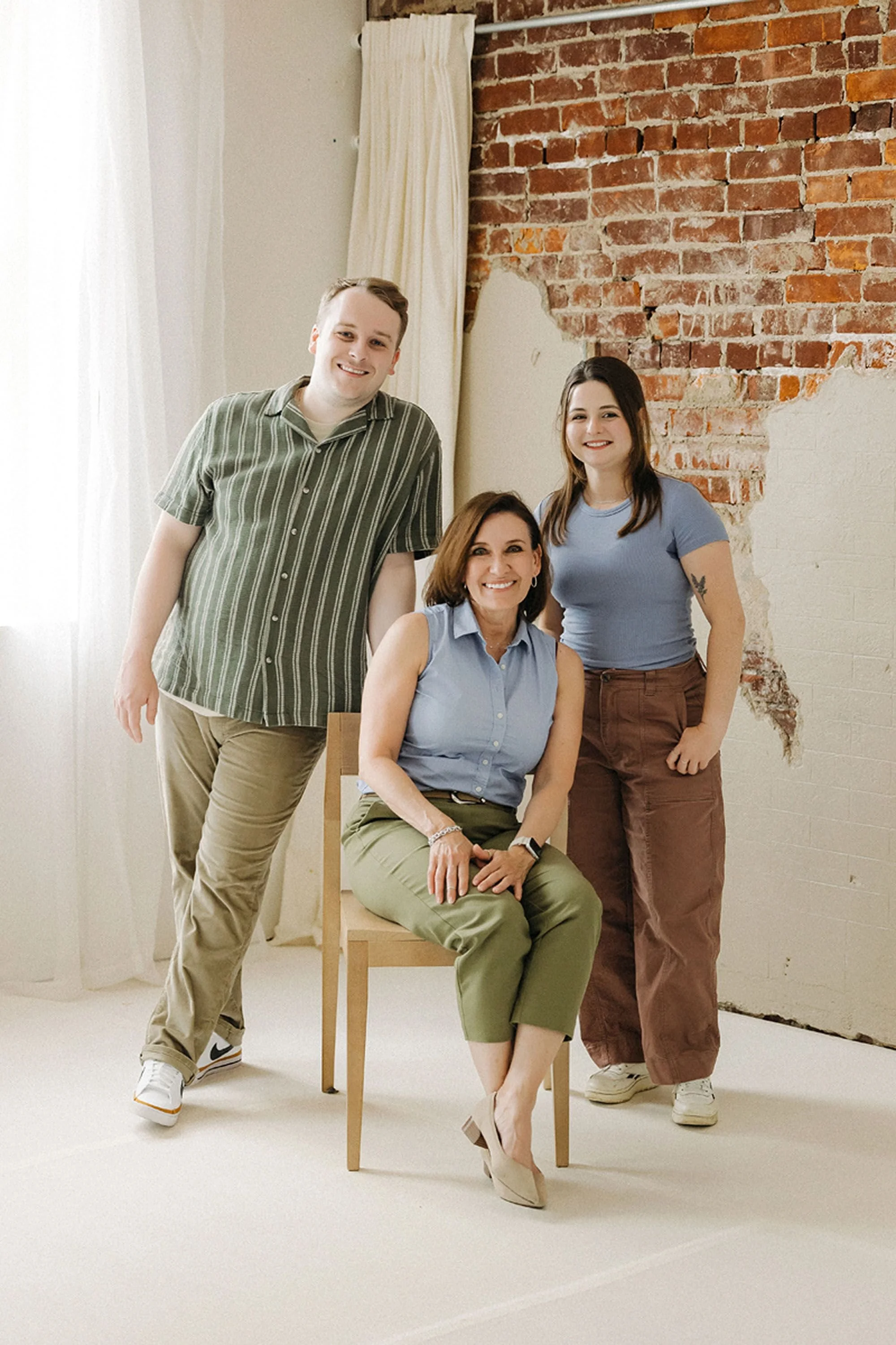 Mother seated with her adult son and daughter standing beside her in a bright loft studio with exposed brick walls, captured during Studio Family Photos at Studio Loft 18