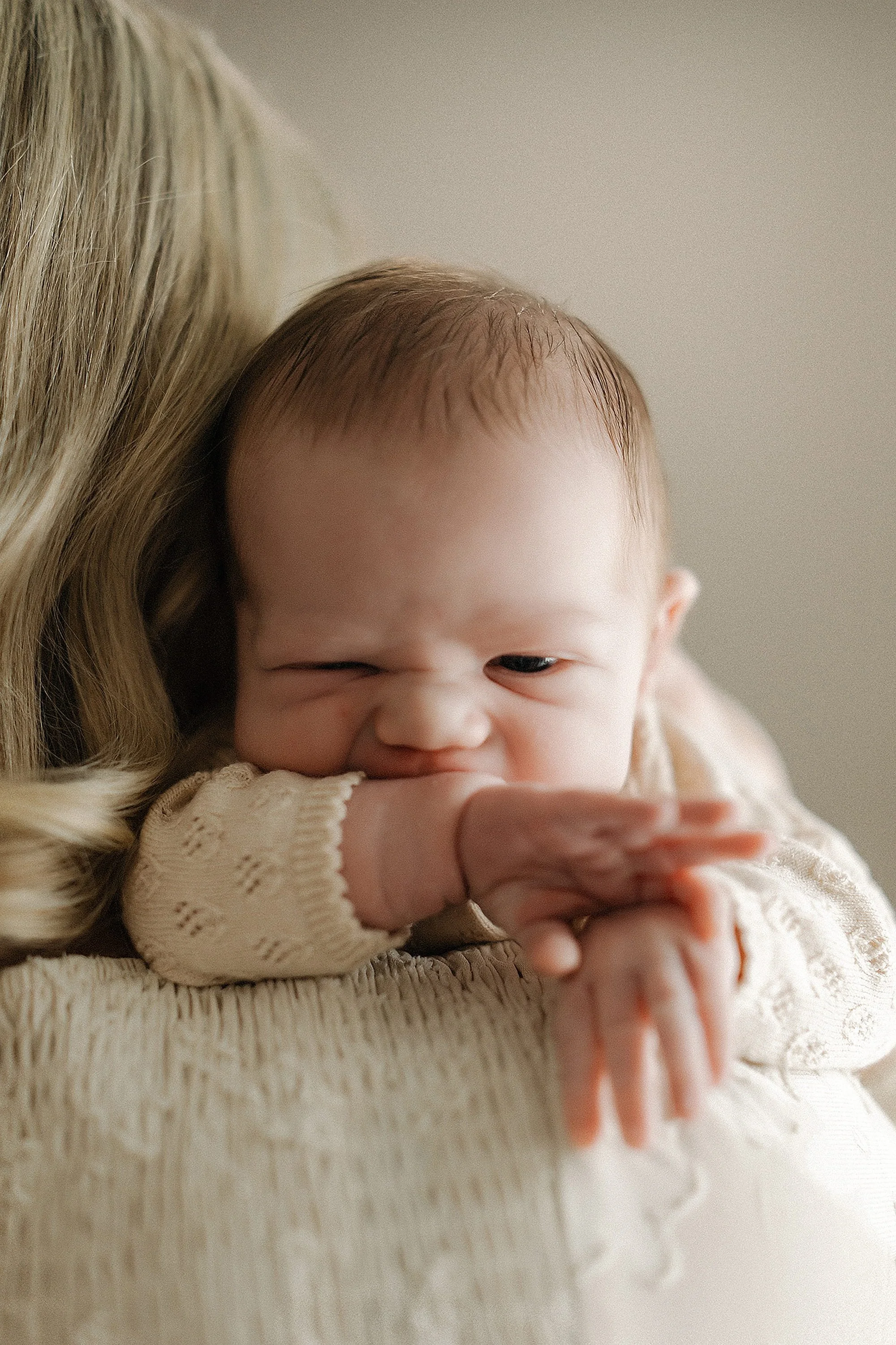 Close-up of a baby girl resting her chin on her arm, lying on an adult's shoulder, with a gentle expression and a cozy beige sweater.