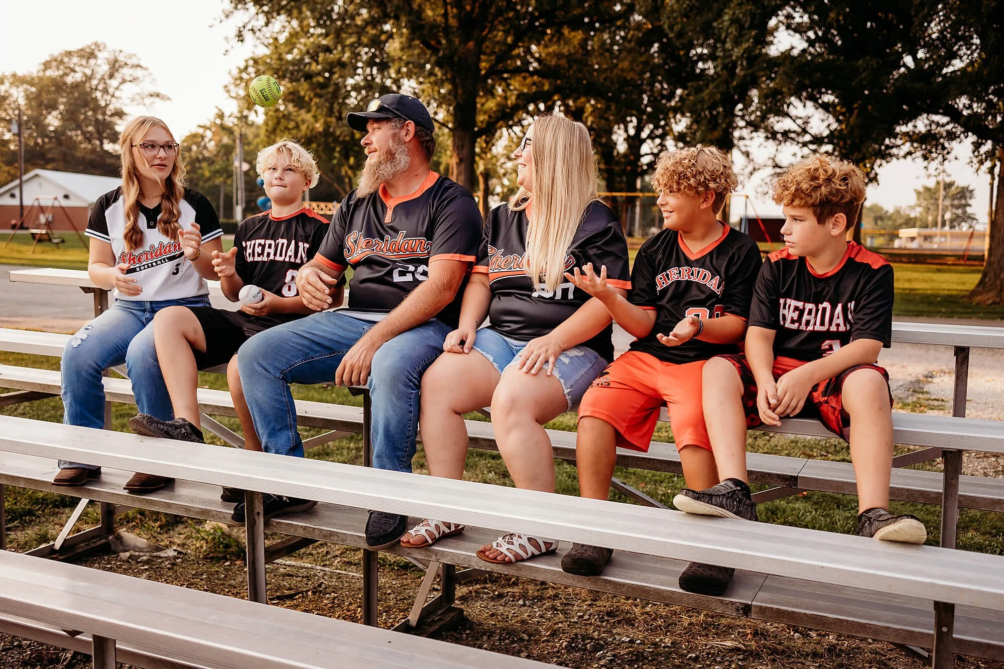 Family sitting on metal bleachers at a baseball field, wearing matching team jerseys, during a playful pregnancy announcement moment with their older kids, captured by theheartnarrative Indiana family photographer