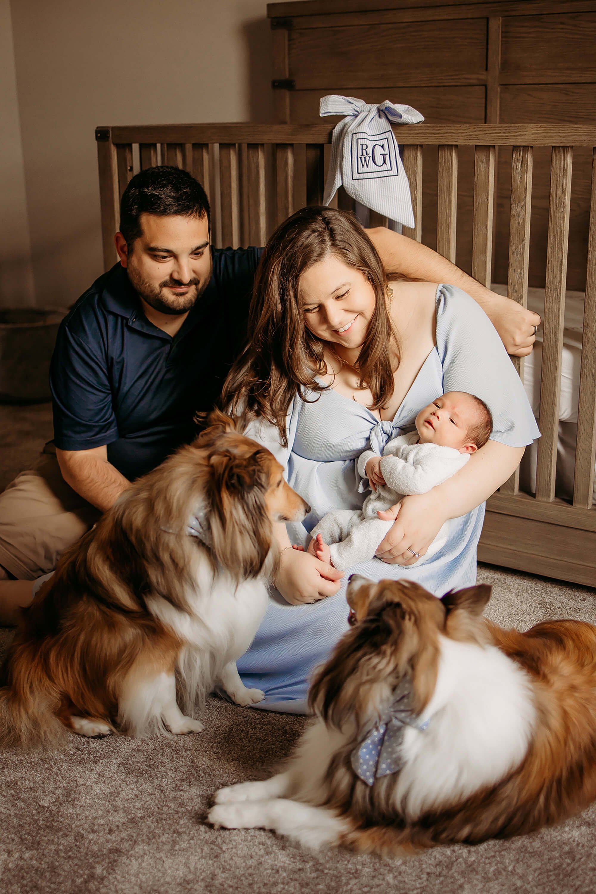new parents, new baby and sheltie dogs lay on floor of nursery, documenting the first year of baby photos in indiana