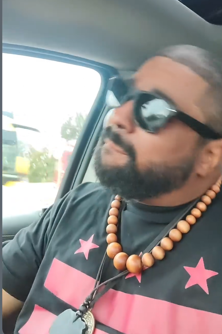 A man with a beard and sunglasses sitting in a vehicle, wearing a black t-shirt with pink stars and a beaded necklace, looking to his left.