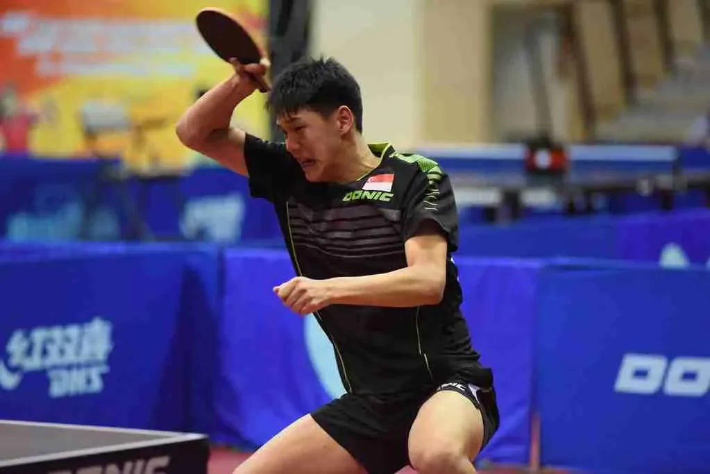 A young man in a black sports shirt and shorts plays table tennis, preparing to hit the ball with a paddle.
