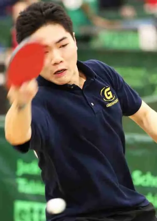 A person playing table tennis, about to hit a white ball with a red paddle, in an indoor sports facility.