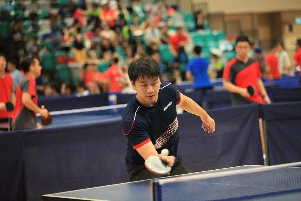 A young man playing table tennis in a crowded indoor space, with spectators and other players in the background.