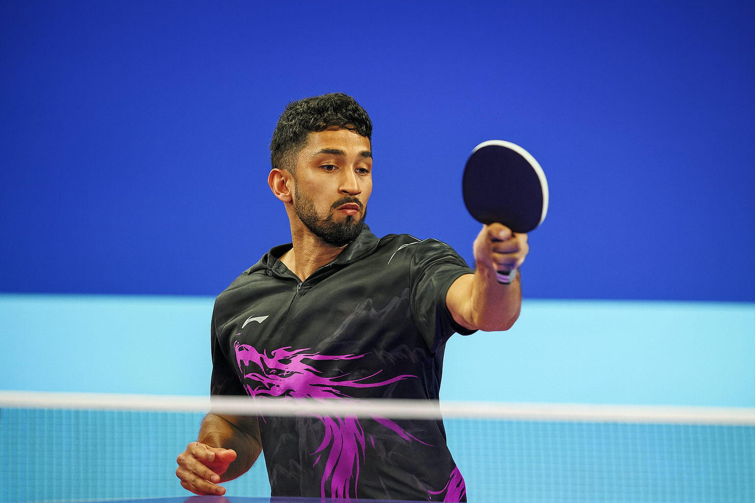 A male table tennis player in a black and purple shirt is playing, holding a paddle in his right hand, with a focused expression.