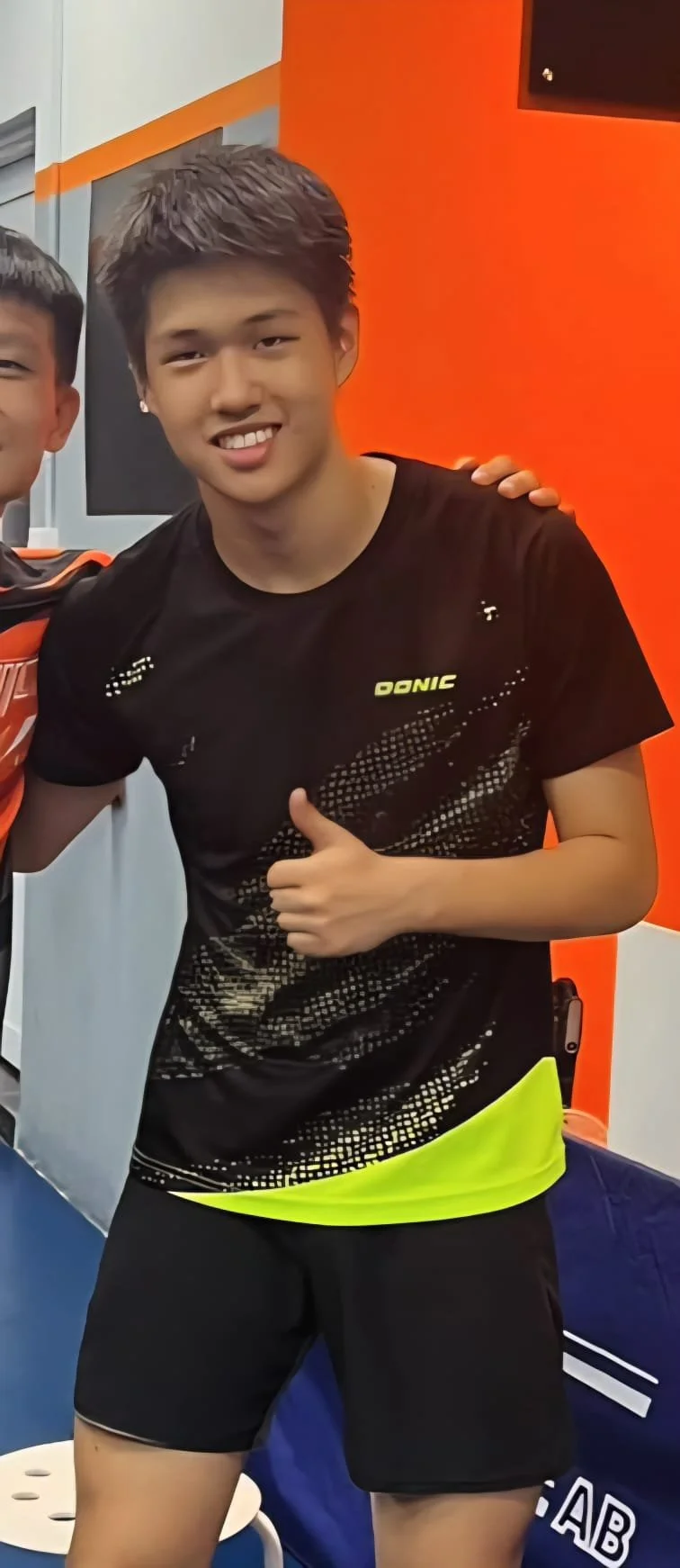 Young man smiling and giving a thumbs-up in sportswear, standing in a sports facility with a bright orange and gray wall behind him.