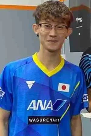 Young man wearing glasses and a blue Japanese sports jersey with the ANA logo, standing indoors.