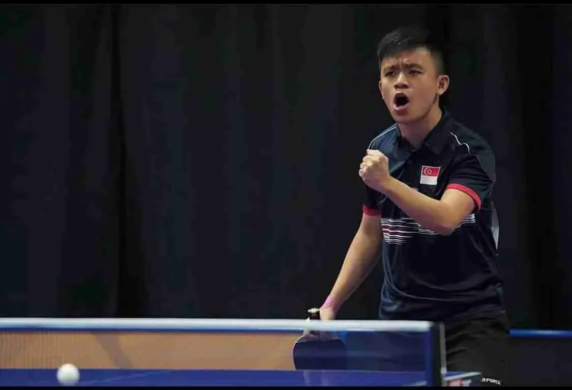 Young man playing table tennis, wearing a black shirt with a Singapore flag patch, making a fist gesture, and expressing enthusiasm or determination.