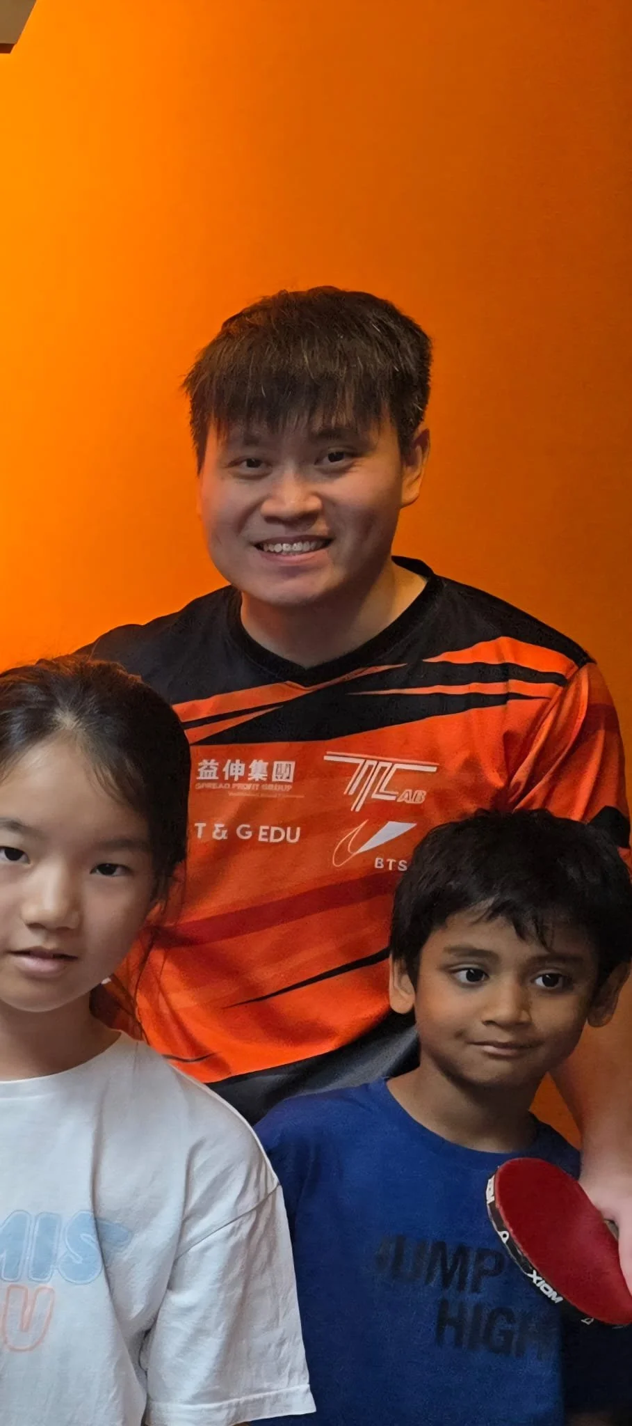 A smiling young man with short dark hair wearing an orange and black sports jersey, standing behind two children, a girl with black hair in a white T-shirt and a boy with black hair in a blue shirt holding a red table tennis paddle. All are against an orange background.