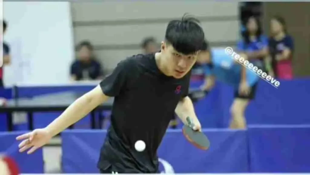 A young man playing table tennis, about to hit the ball with his paddle, in an indoor gym with other players in the background.