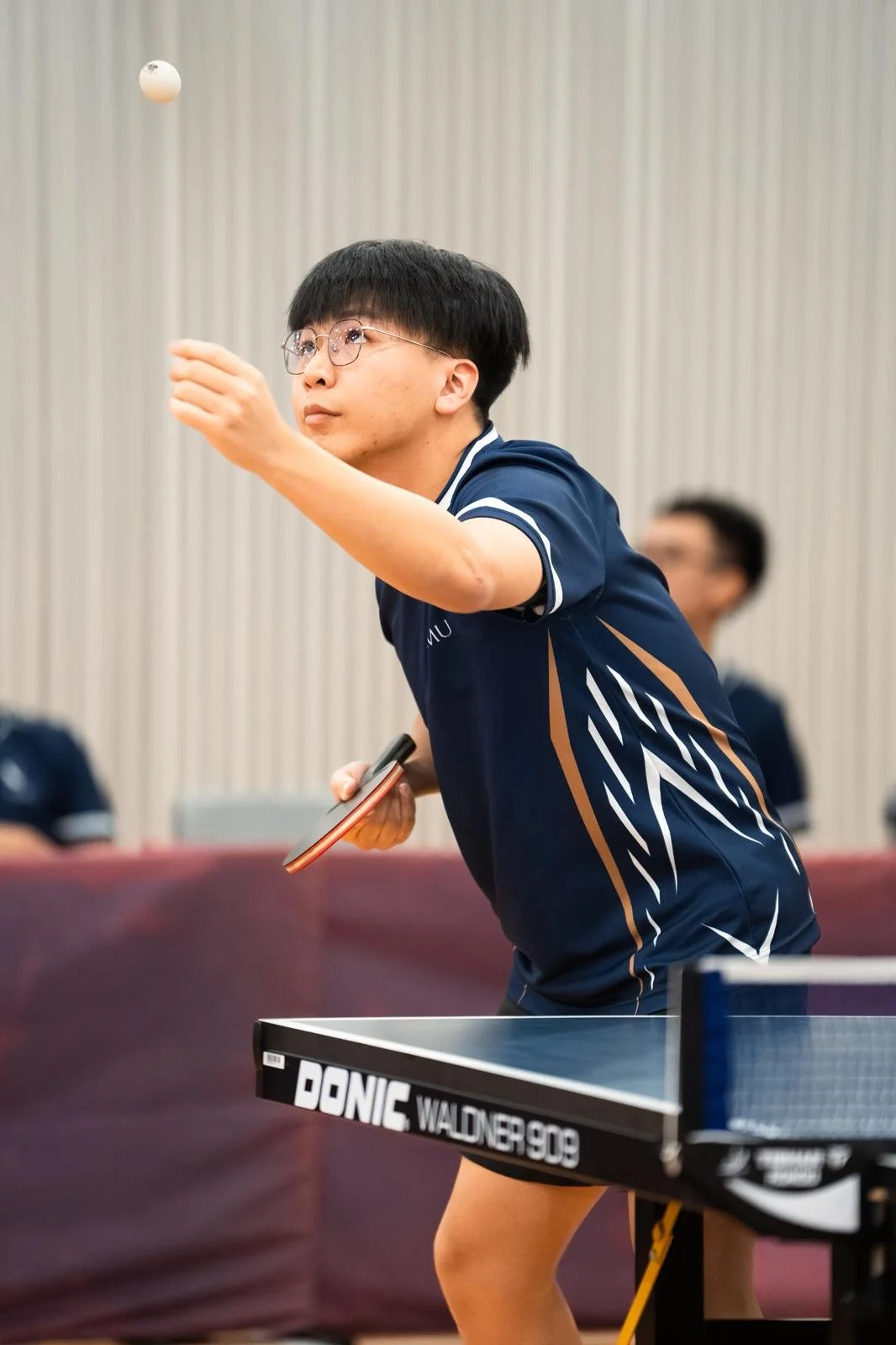 A young man with glasses playing table tennis, holding a paddle in one hand and preparing to hit a white ping pong ball.