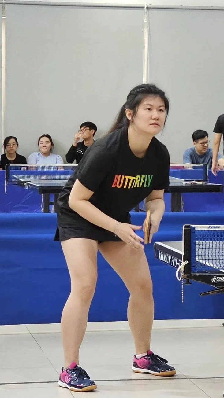 Young woman in black T-shirt with 'BUTTRFLY' in rainbow colors, black shorts, and sneakers playing table tennis in an indoor gym.