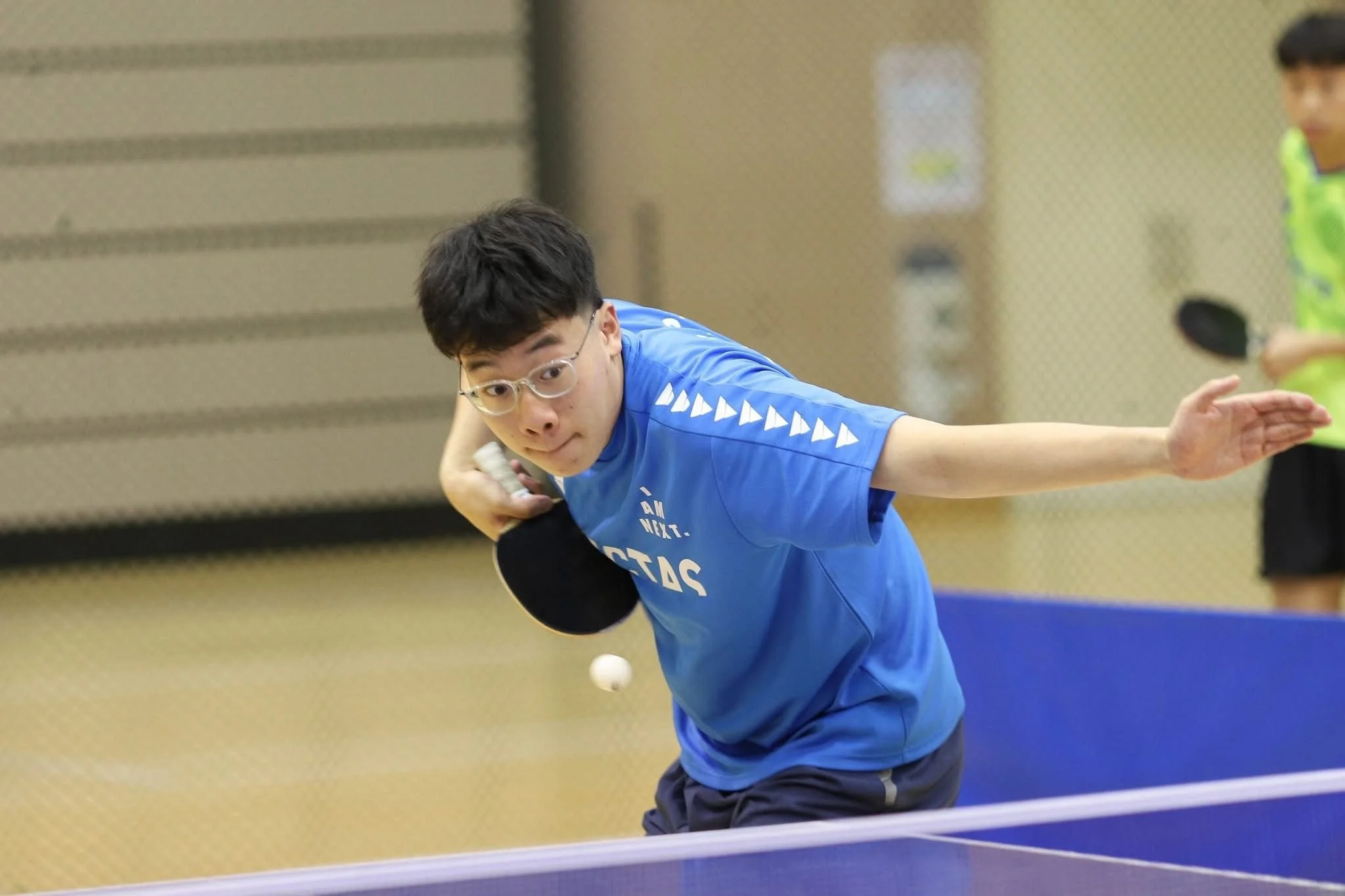 A young man in glasses wearing a blue t-shirt and shorts playing table tennis, about to hit the ball.
