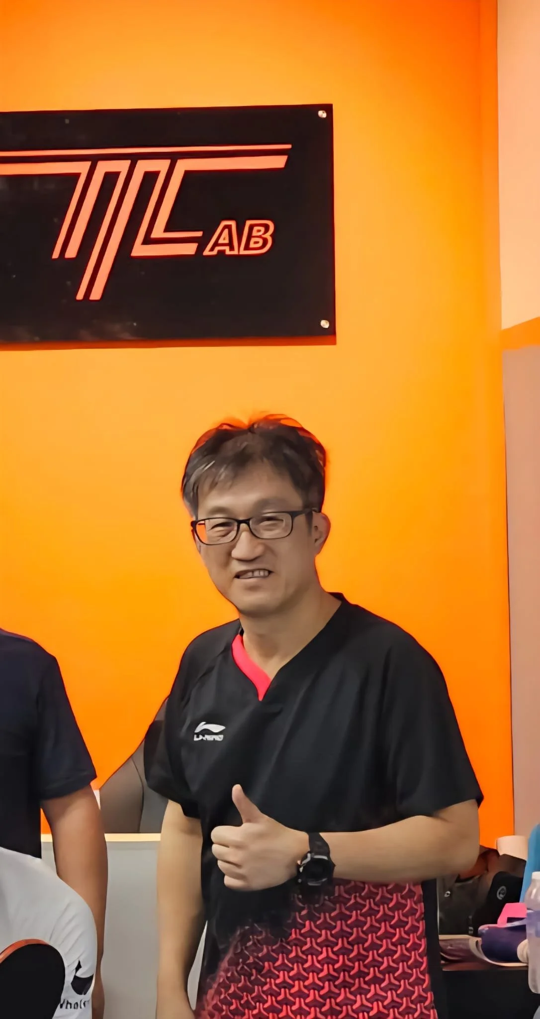 A man with glasses giving a thumbs-up in front of an orange wall with a black and red logo and sign reading 'TTC AB'
