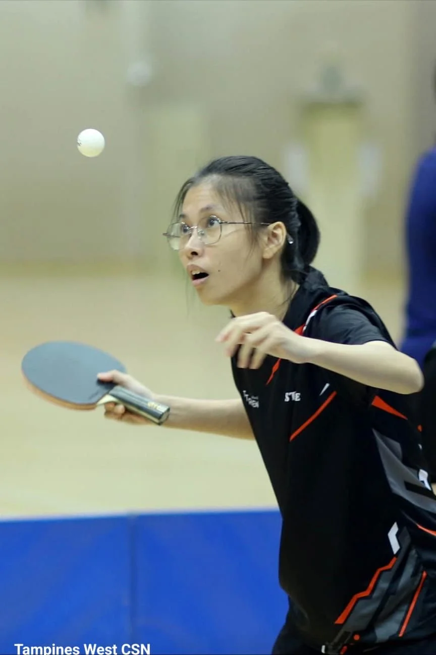 A woman with glasses playing table tennis, focused on the ball, in an indoor gym with blue and tan walls.