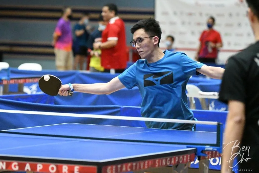 A young man in a blue T-shirt and glasses playing table tennis, hitting the ball with a black paddle at an indoor table tennis facility.