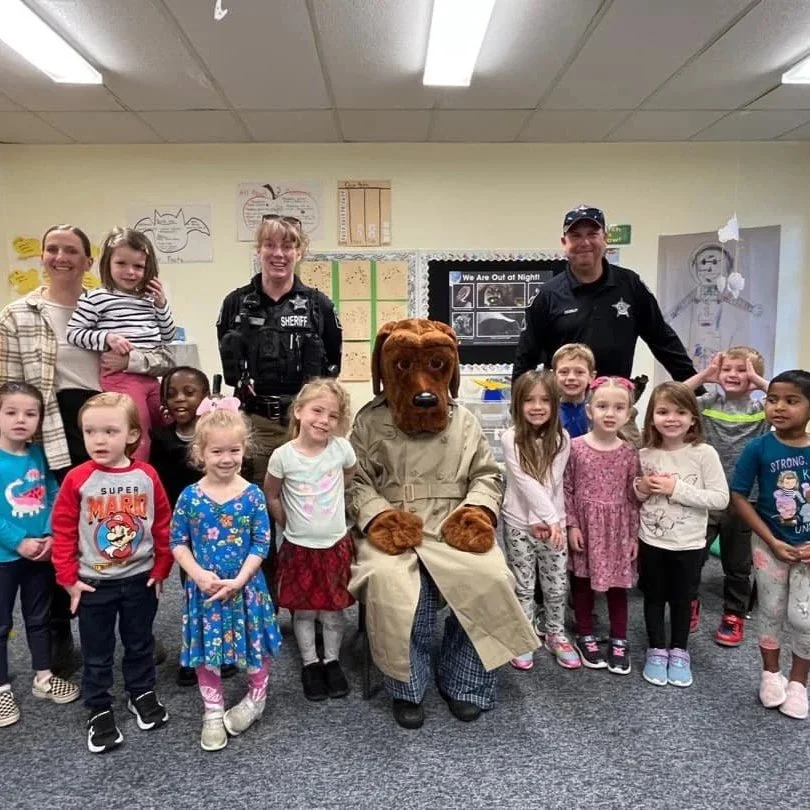 Group of children, police officers, and a person in a dog costume in a classroom.