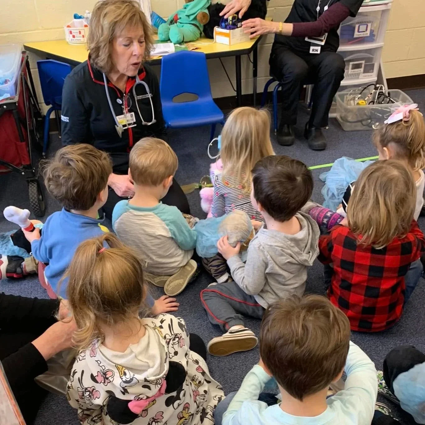 A group of young children sitting on the floor of a classroom listening to a nurse who is talking to them. The nurse is wearing a black shirt with a stethoscope and has a badge around her neck. In the background, there are toys and medical supplies o