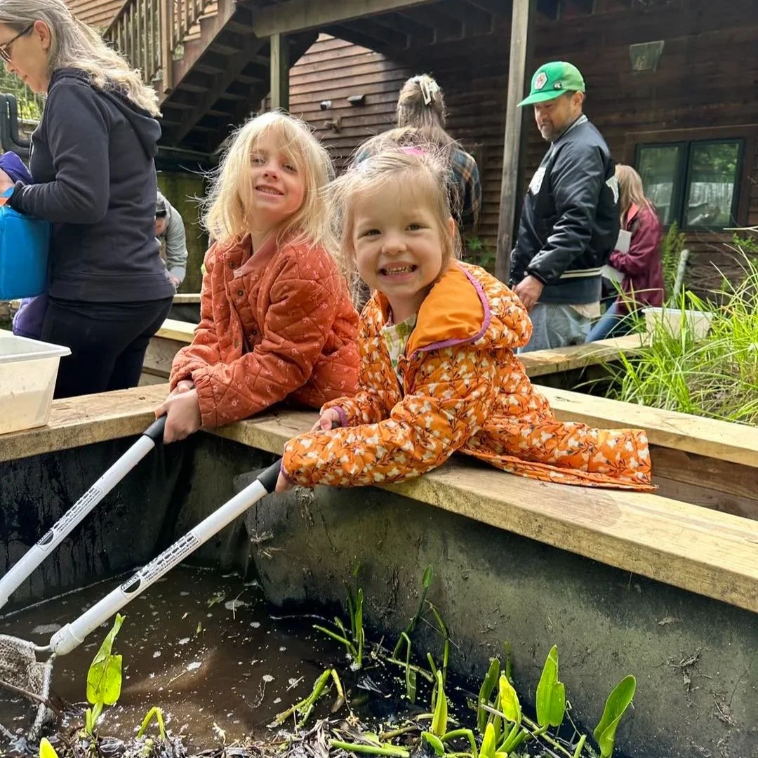 Two young girls are leaning over the edge of a pond with butterfly nets, smiling at the camera, with adults and a house in the background.