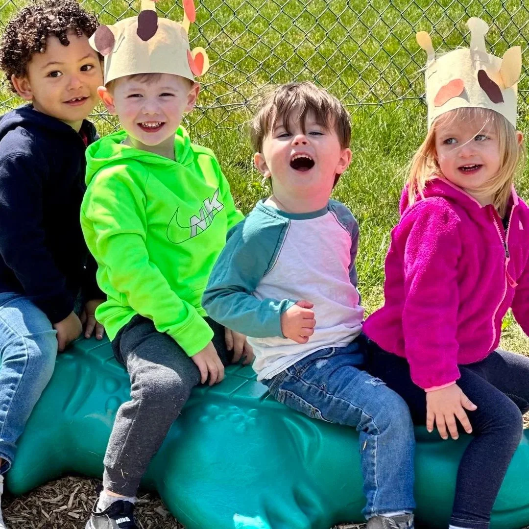 Four children sitting on a green dinosaur-shaped playground sculpture, wearing animal-themed paper crowns, outside near a chain-link fence under bright daylight.