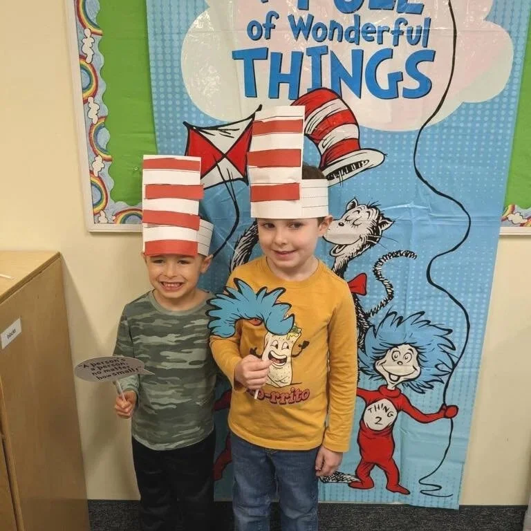 Two children wearing striped red and white hats inspired by the Dr. Seuss book 'The Cat in the Hat'. They are standing in front of a blue Dr. Seuss themed backdrop with the title 'The Pool of Wonderful Things'. One child is holding a paper fan with t