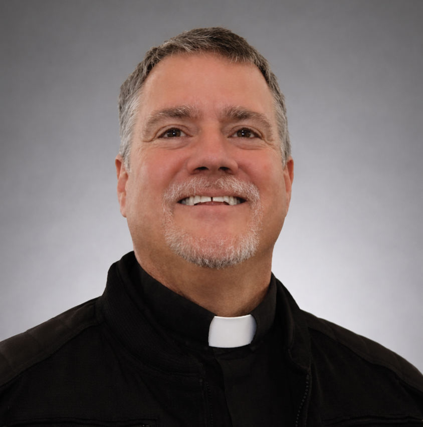 Portrait of a smiling middle-aged man wearing glasses, a clerical collar, and a dark blazer against a gray background.