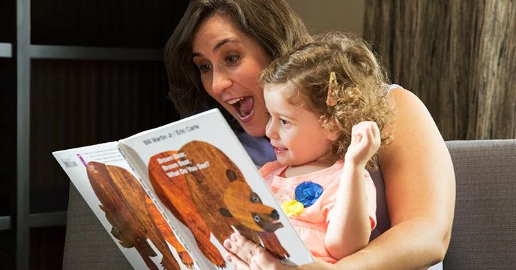 A woman reading a children's book to a young girl, both smiling and enjoying the story together.
