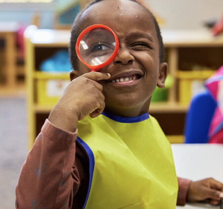 Young boy holding a magnifying glass up to his eye, smiling in a classroom setting.