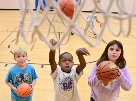 Children playing basketball in a gym, with one boy shooting a basketball towards the hoop.
