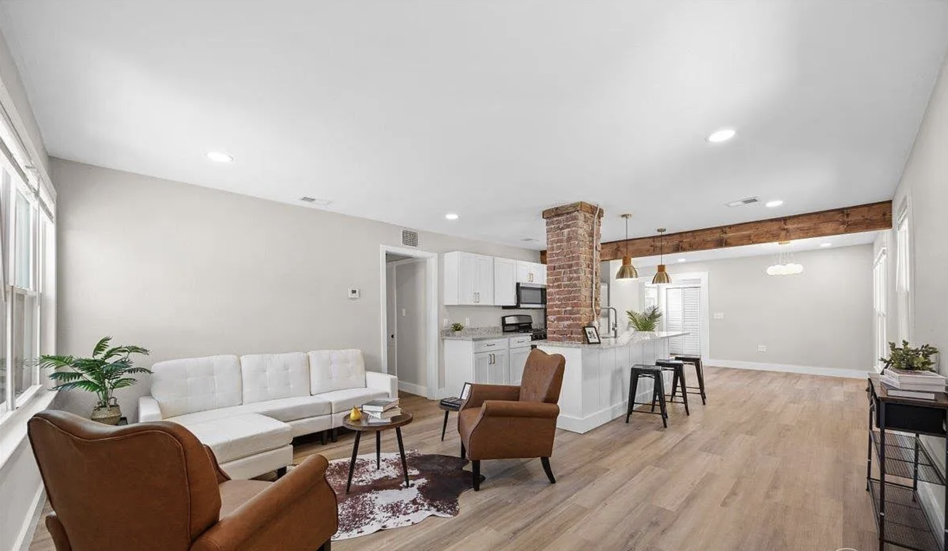 Open-concept living room and kitchen with white walls, wooden flooring, a brick column, white cabinets, bar seating, and modern furnishings.