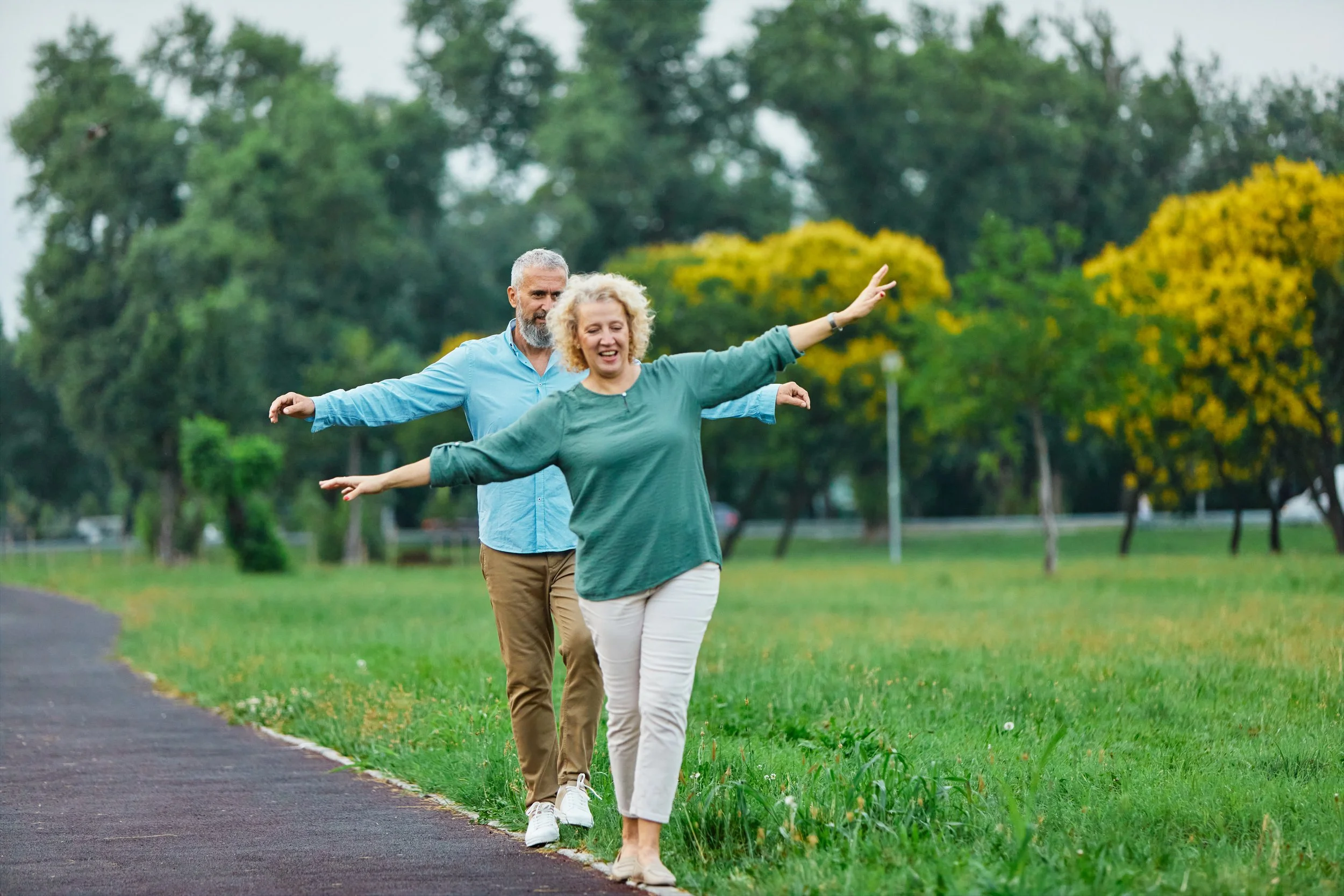 A man and woman walking and balancing on a narrow grassy border beside a paved path in a park, with the woman smiling and raising her arms, and the man following behind with his arms slightly outstretched, surrounded by green trees and yellow flowers.