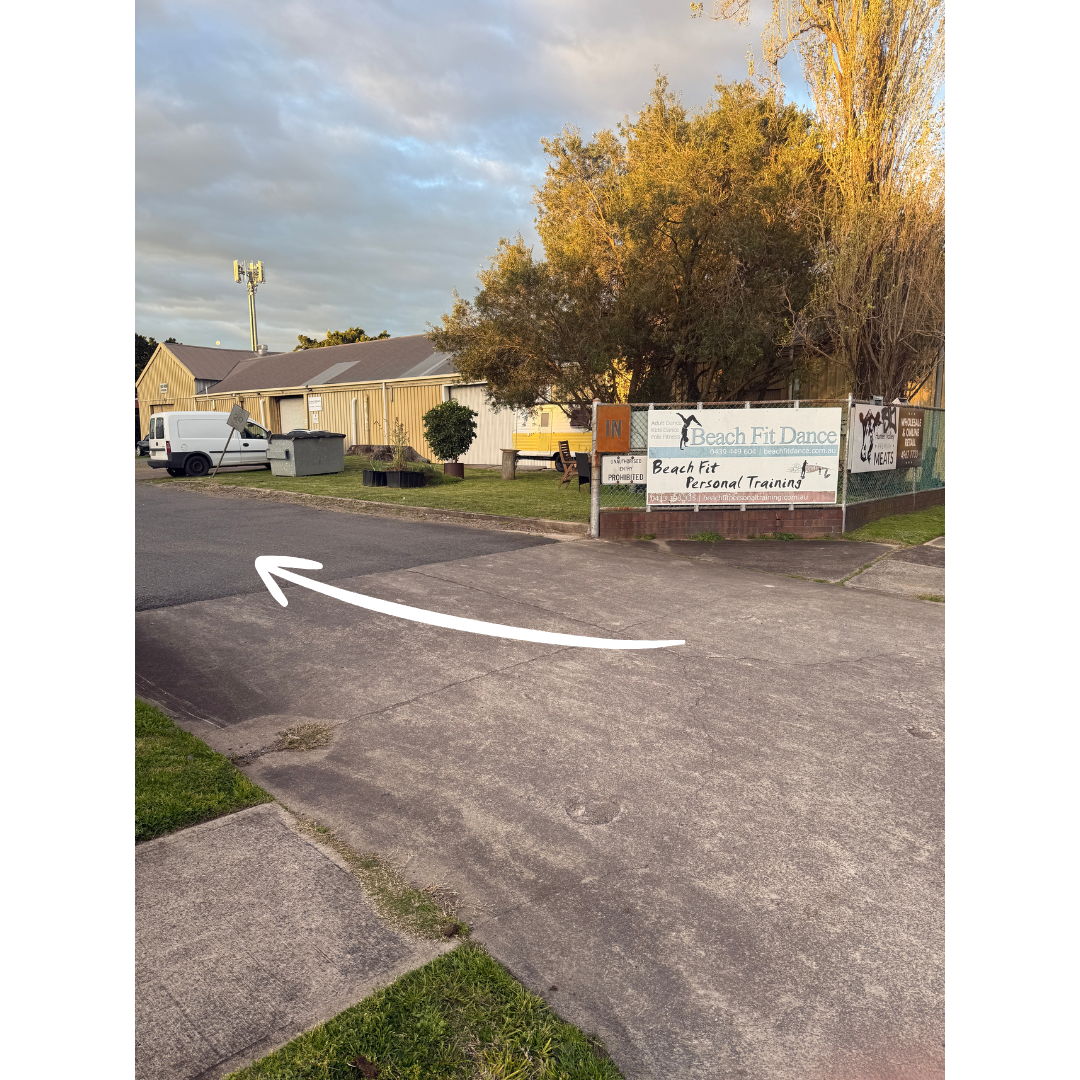 A sidewalk with an arrow painted on it pointing left, leading to a parking lot next to a street. In the background, there's a small building with signs advertising Beach Fit Dance and personal training, surrounded by trees and residential buildings.