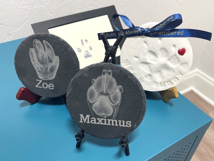An arrangement of stone and clay paw prints, two of which have the name of the pet lasered onto the stone. The clay paw print has a personalized blue ribbon attatched to it.