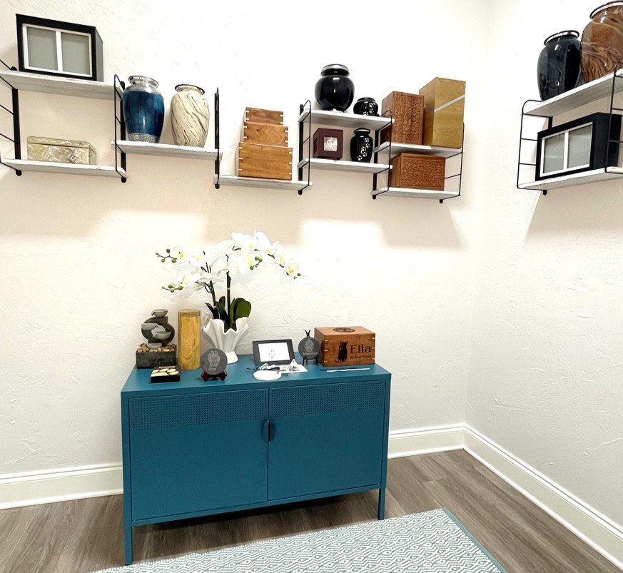 A beautifully appointed room with different styles of urns for pets. A teal cabinet is shown with a orchid on top next to stone, paper, and clay paw prints.