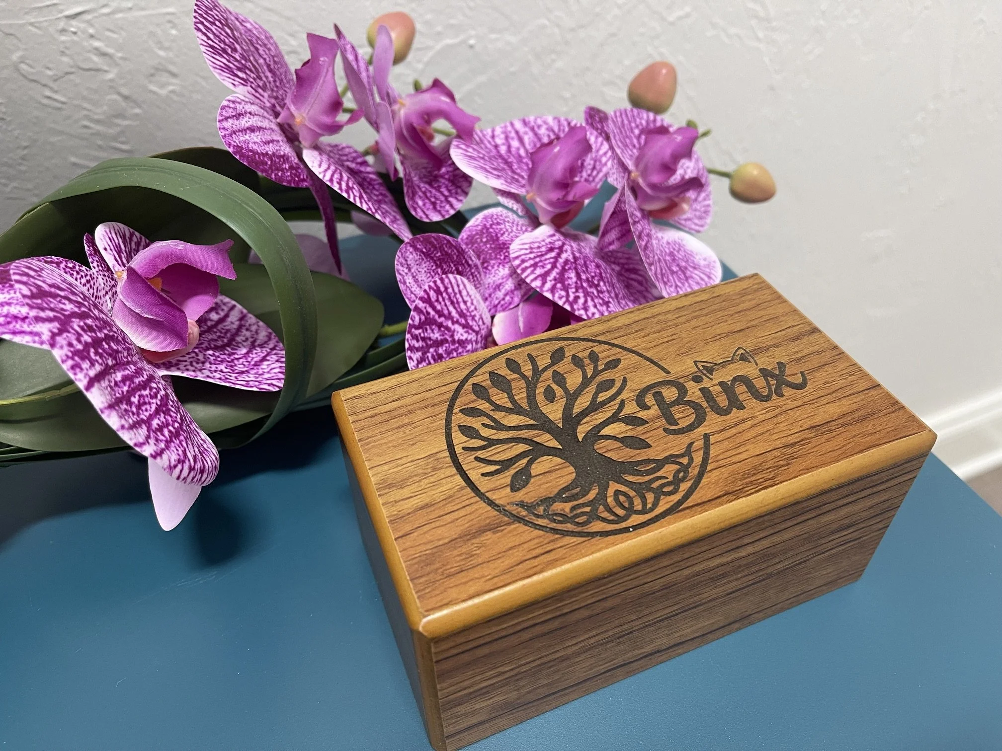 A brown wood urn engraved with the tree of life and the name "Binx" next to it with a set of cat ears over the letter "n". The urn sits on a blue table next to a pink orchid.