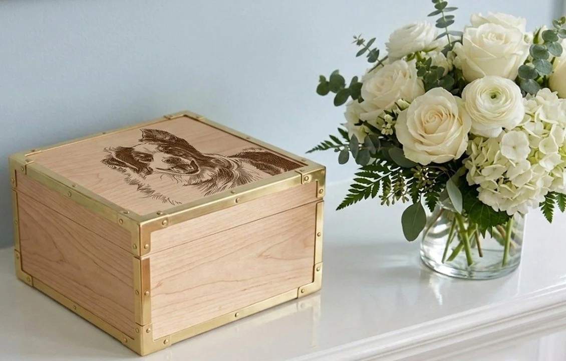 A wood urn with a custom engraving of a dog on the top. The urn has gold trim and is sitting on a white sgelf with a bouquet of white roses next to it.