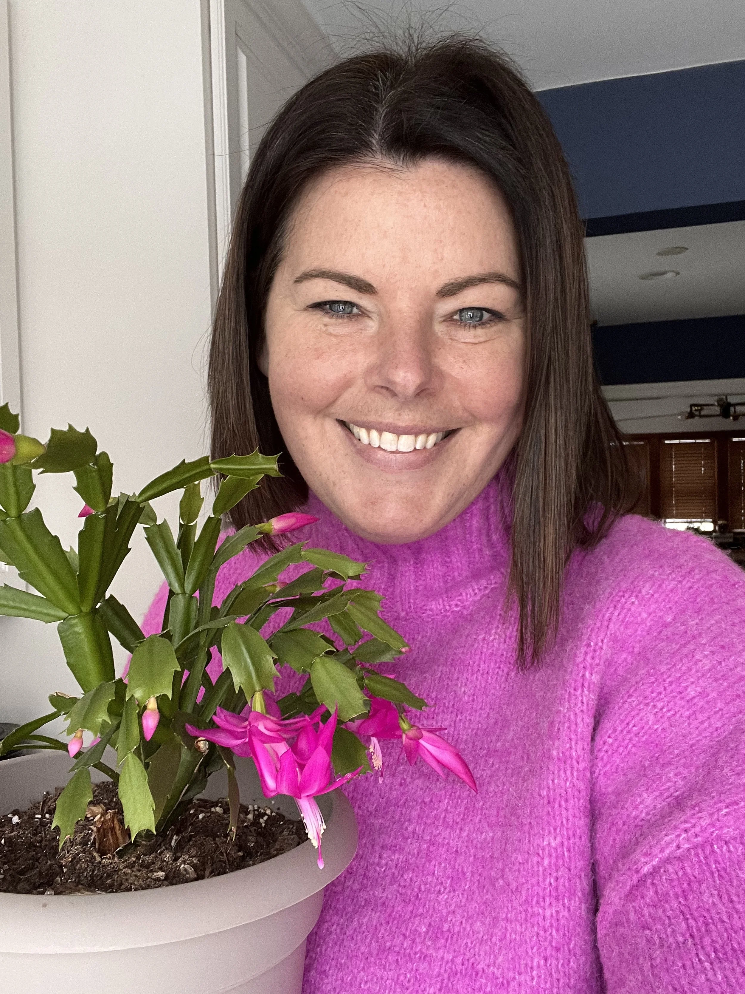 A woman with shoulder-length brown hair and blue eyes smiling, wearing a pink sweater, next to a potted pink flowering plant inside a home.