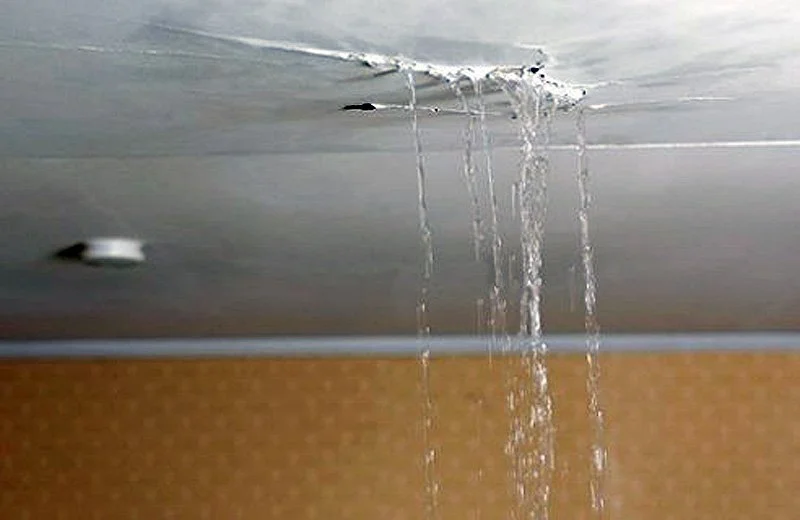 Water leaking from a ceiling with a hole, causing water to drip down, above a textured wall.