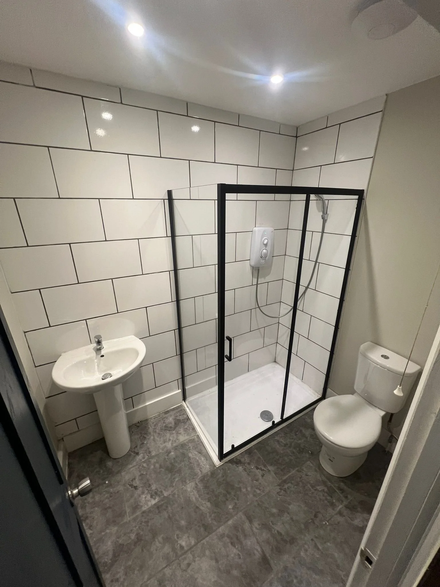 Small bathroom with white tiled walls, a pedestal sink, a toilet, and a walk-in shower with a black frame and white shower base.