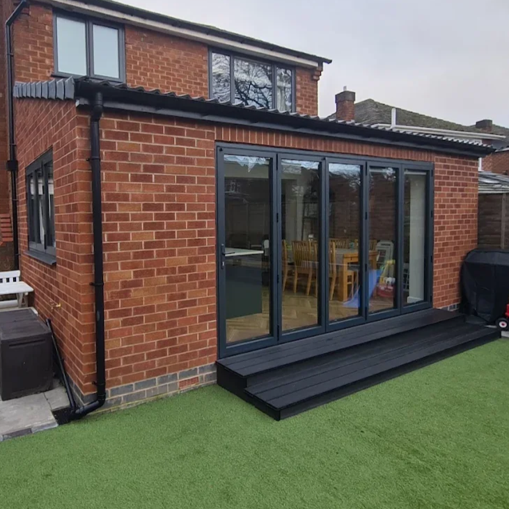 Rear view of a brick house with a modern glass sliding door leading to a small patio with black decking, a green lawn, and a barbecue grill on the side.