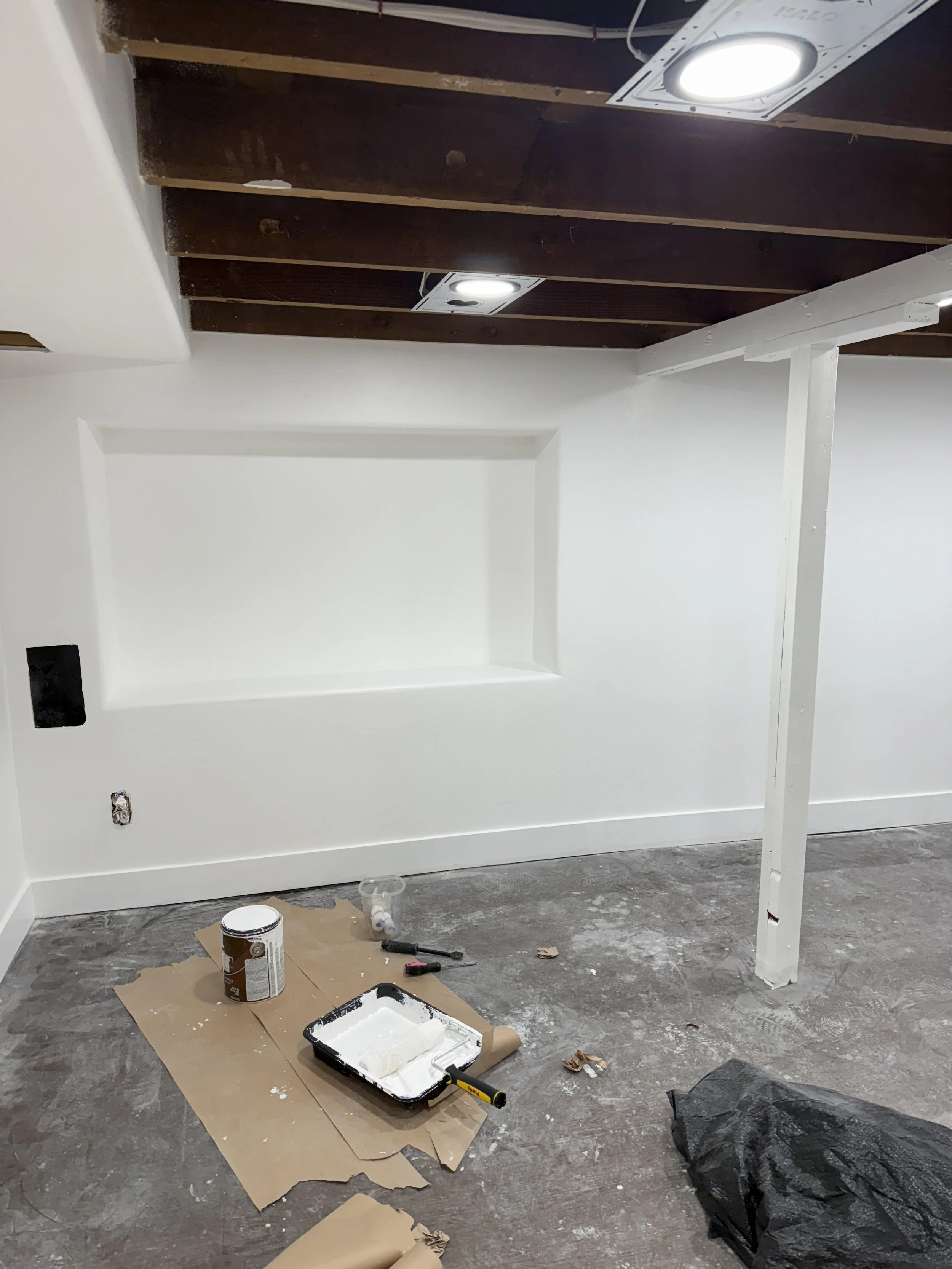 Room under renovation with white painted wall, exposed dark wooden ceiling beams, ceiling lights, and construction tools on the floor, including a paint roller, paint tray with white paint, and a black plastic sheet.