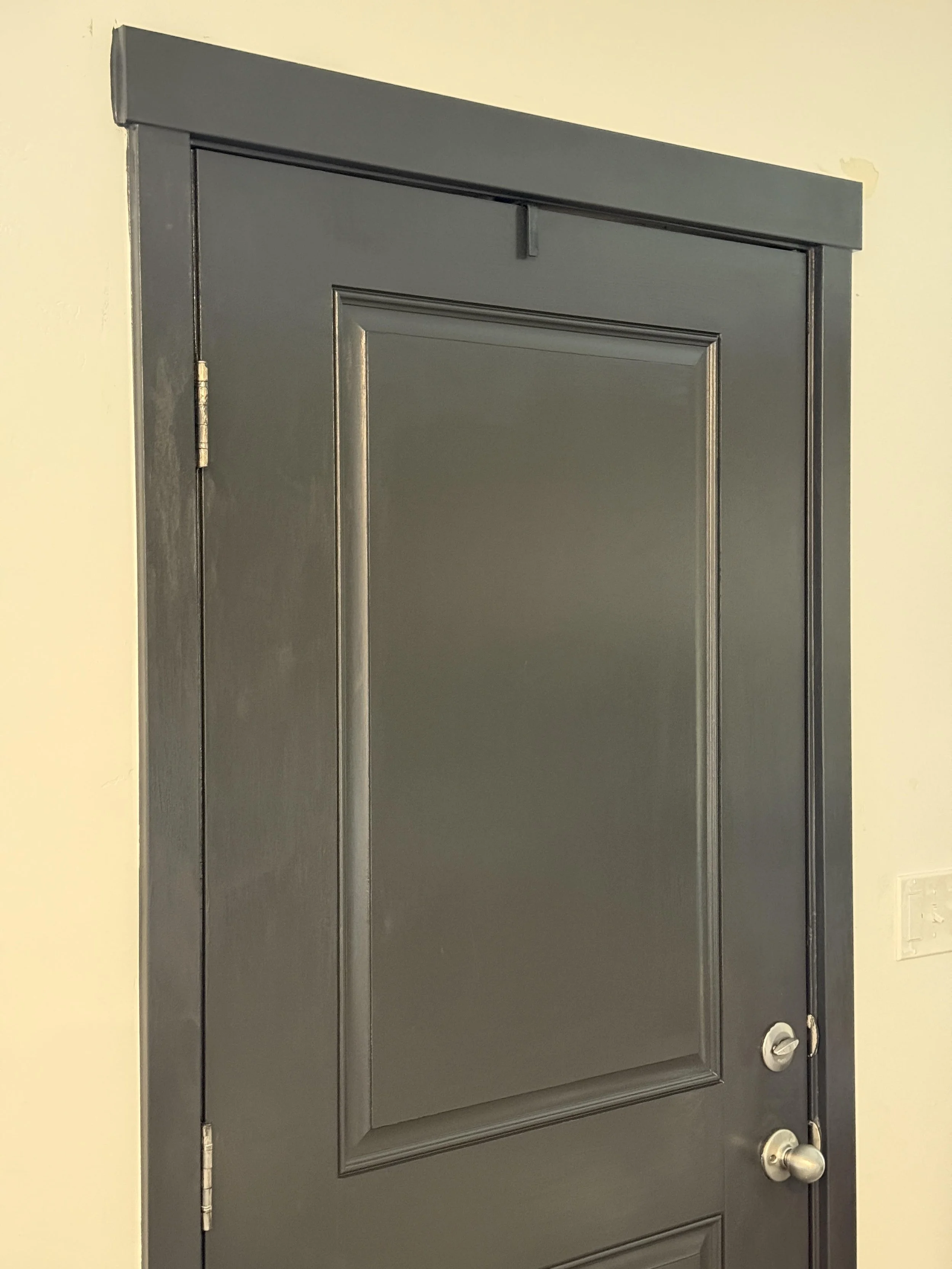A dark gray front door with a silver doorknob and deadbolt, surrounded by a matching gray door frame, and a small gap at the top where the door meets the frame, set in a beige wall.