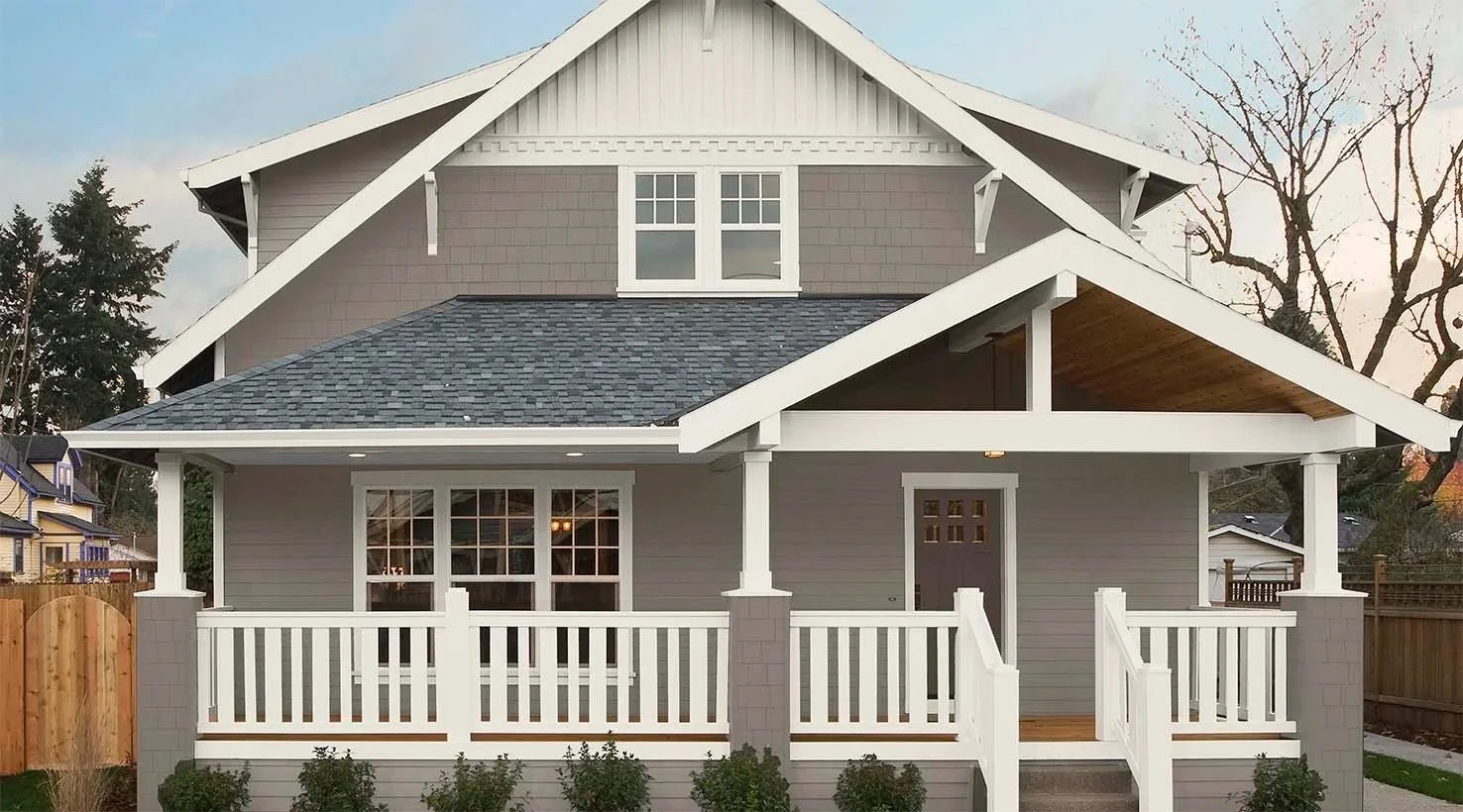 Front view of a two-story gray house with a large front porch, white railings, and a gabled roof.