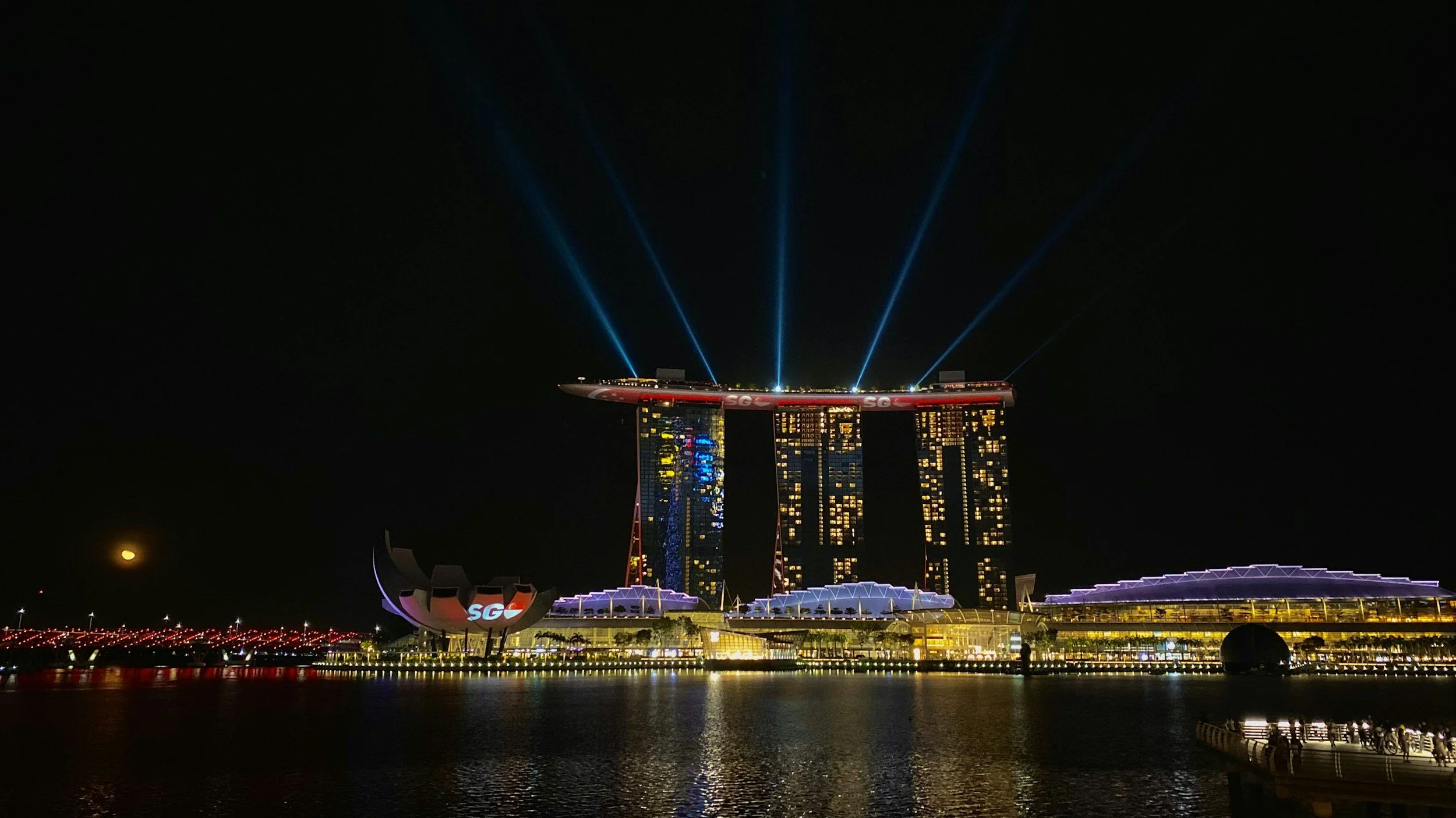 Night view of Marina Bay Sands hotel in Singapore with laser lights on top, reflecting on the water below.
