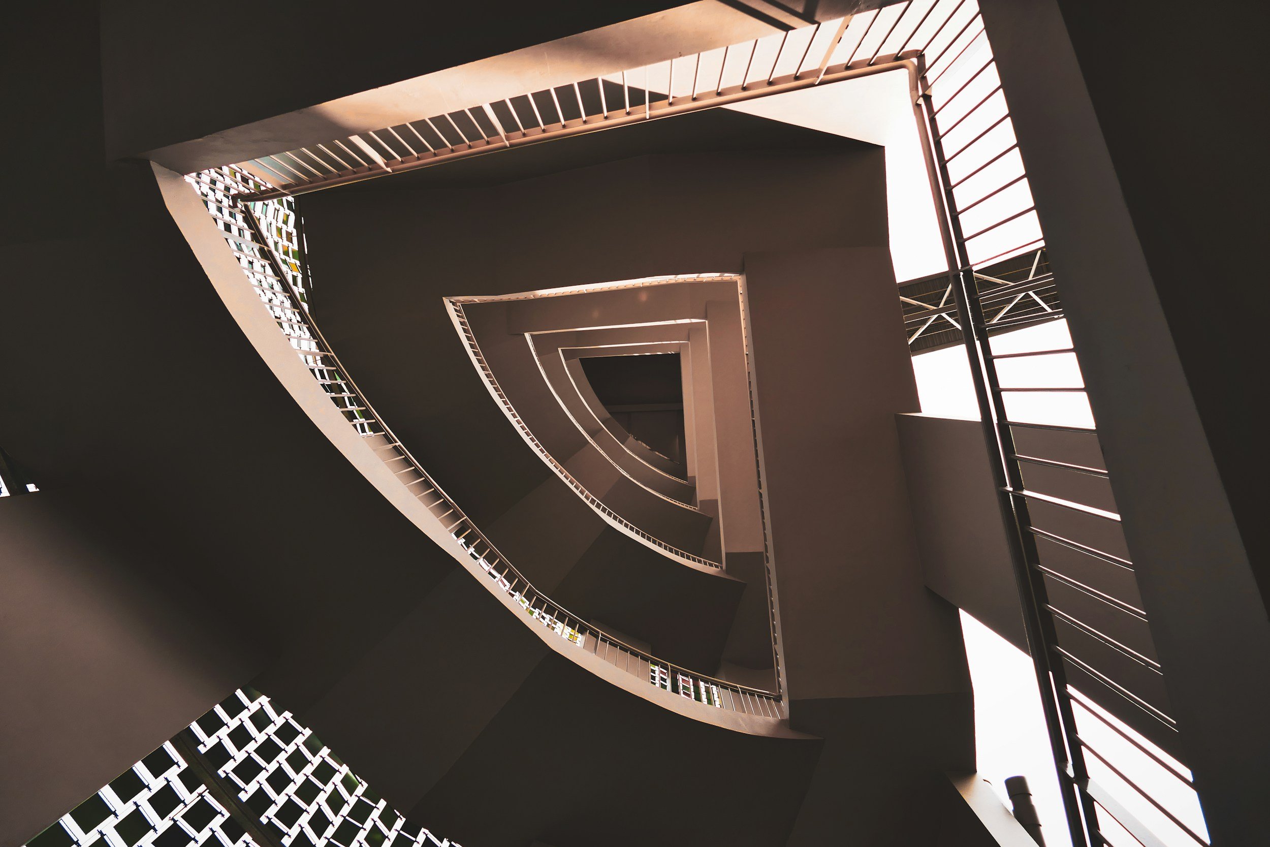 Looking up a spiraling staircase from the bottom, showing multiple levels with railings, open ceiling with natural light, and geometric architectural design.