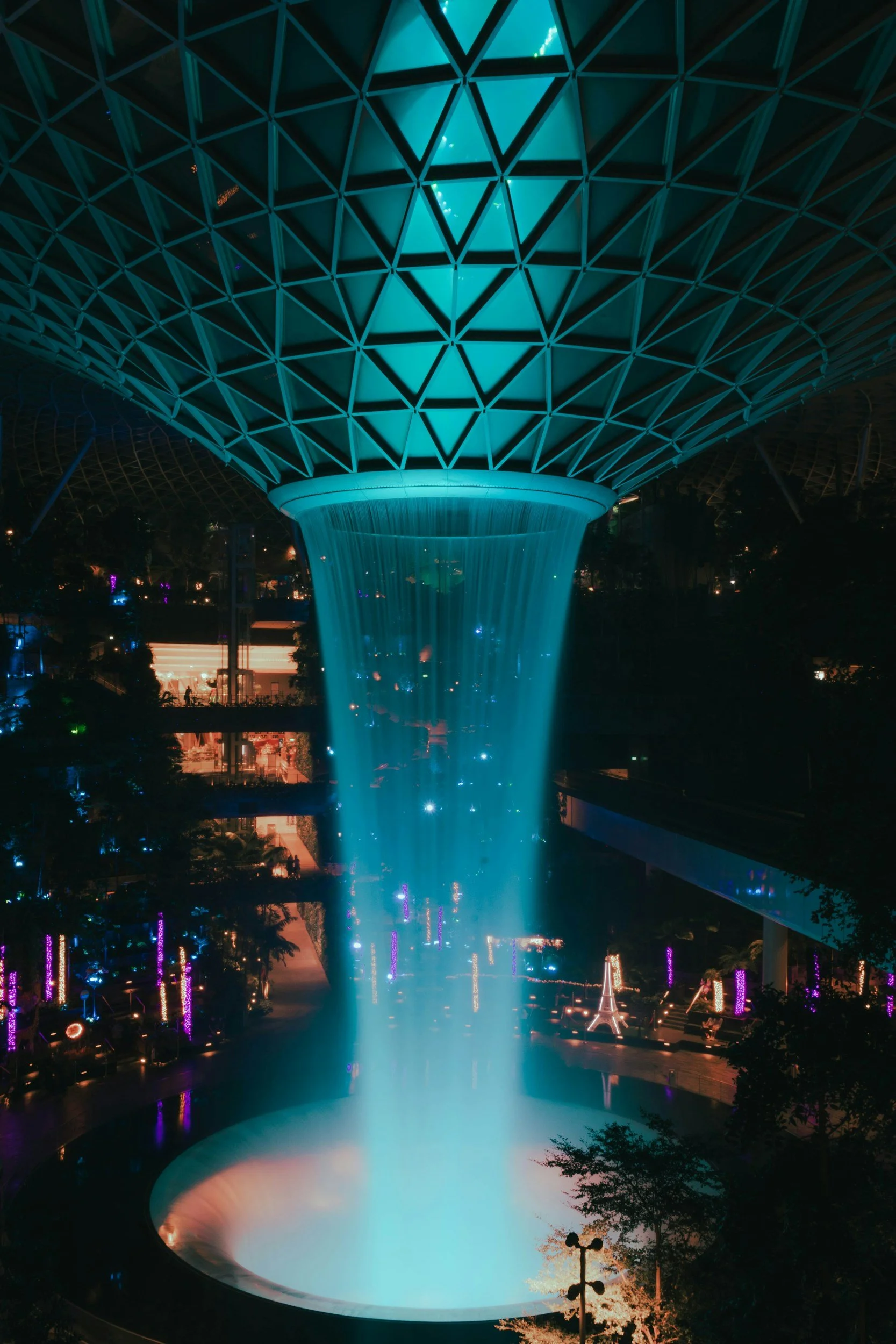Night scene of a large, illuminated indoor waterfall with a geometric, dome-shaped ceiling. The waterfall appears to flow from the ceiling into a circular pool below, with colorful lighting creating a vibrant atmosphere.