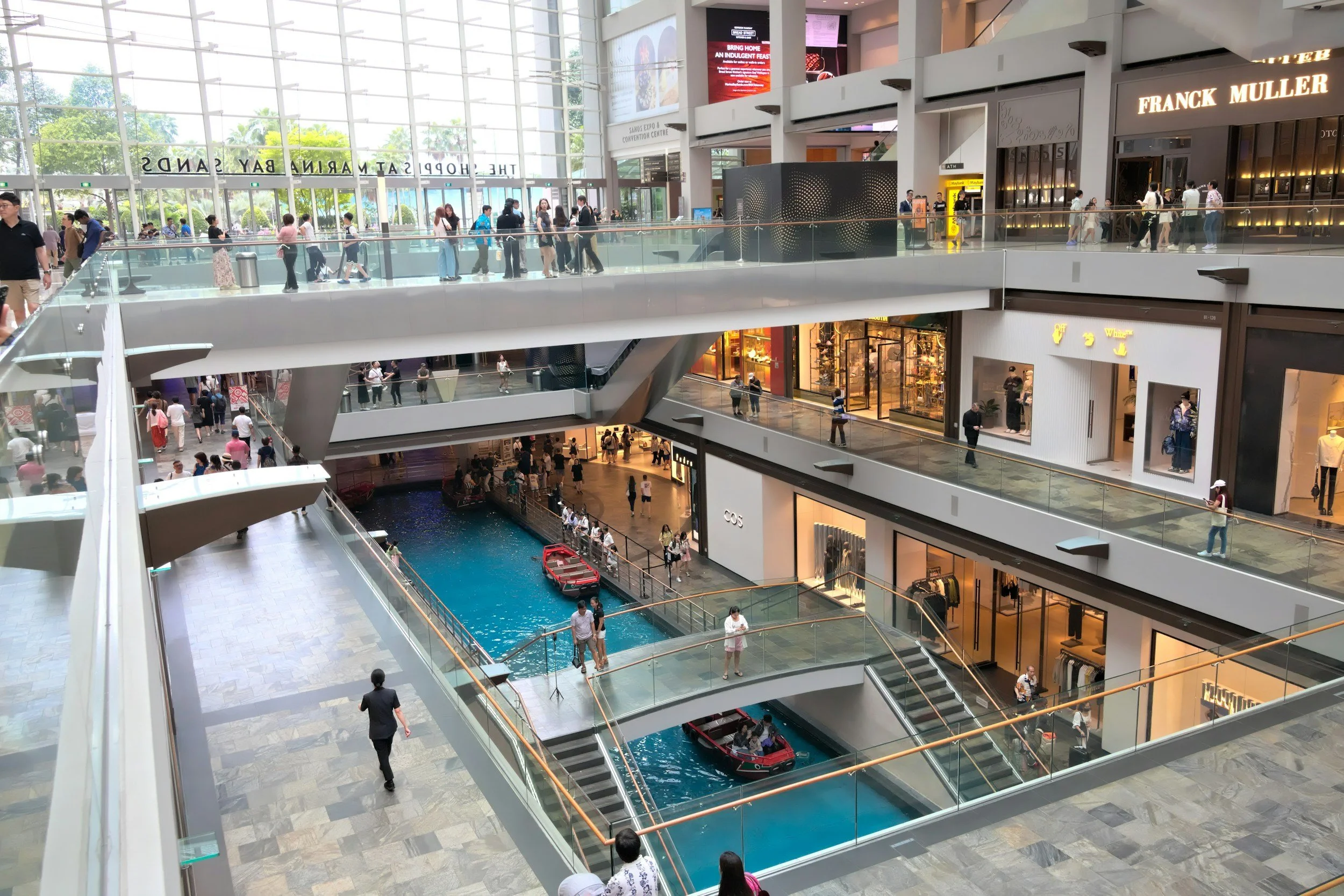 Interior view of a modern shopping mall with multiple floors, glass railings, and shops. A water feature with boats is visible on the lower level, and people are walking on all levels.