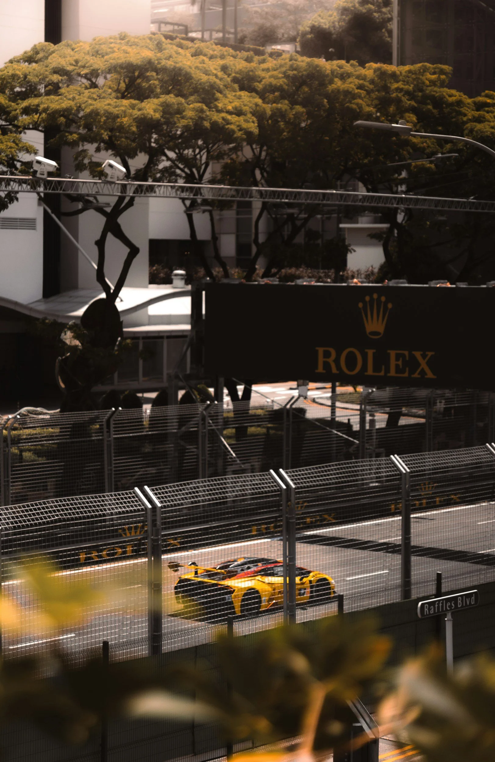 A yellow race car is driving on a racetrack behind a safety fence, with trees and modern buildings in the background. The track has signage with the Rolex logo and the name Raffles Blvd.